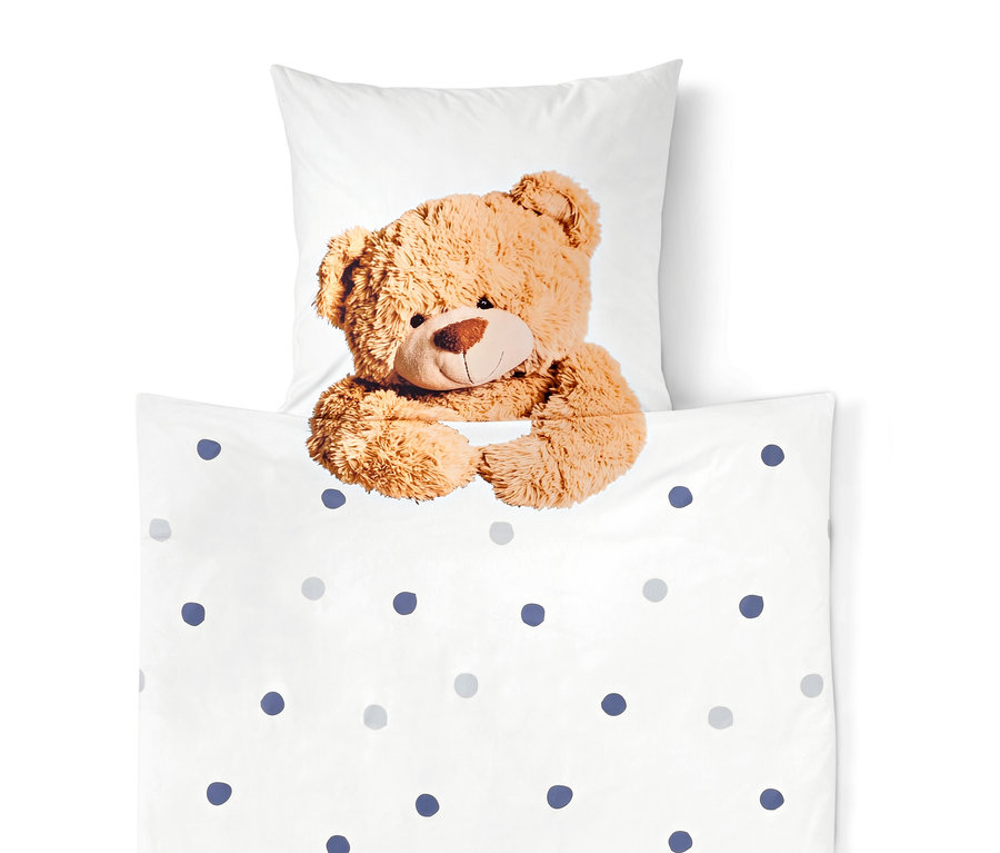 Brązowy miś wygląda zza białej kołdry w niebieskie i szare kropki, w tle biała poduszka.