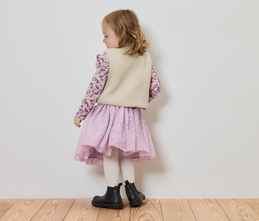 Mała dziewczynka stoi tyłem do kamery, ubrana w bluzkę w kwiaty, jasną kamizelkę, liliową spódniczkę i czarne botki.