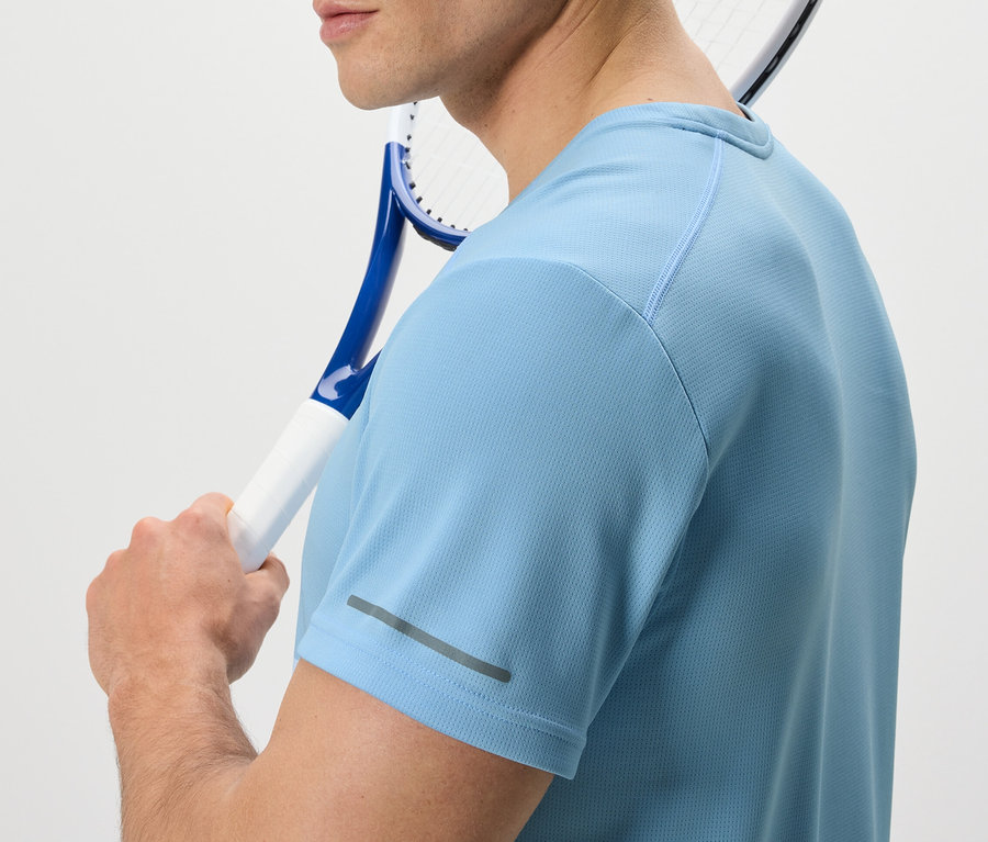 Mężczyzna z rakietą tenisową przewieszoną przez ramię ma na sobie jasnoniebieską koszulkę funkcyjną.