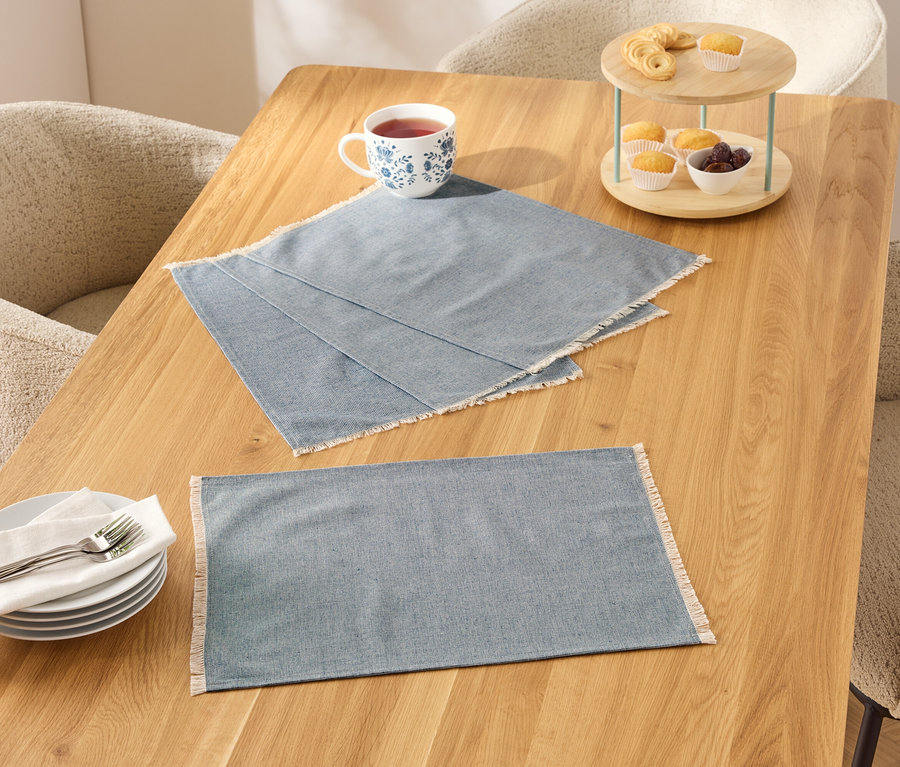 Dwie niebieskie podkładki na stół z białym brzegiem na drewnianym stole, obok nich filiżanka, talerze i etażerka z jedzeniem.