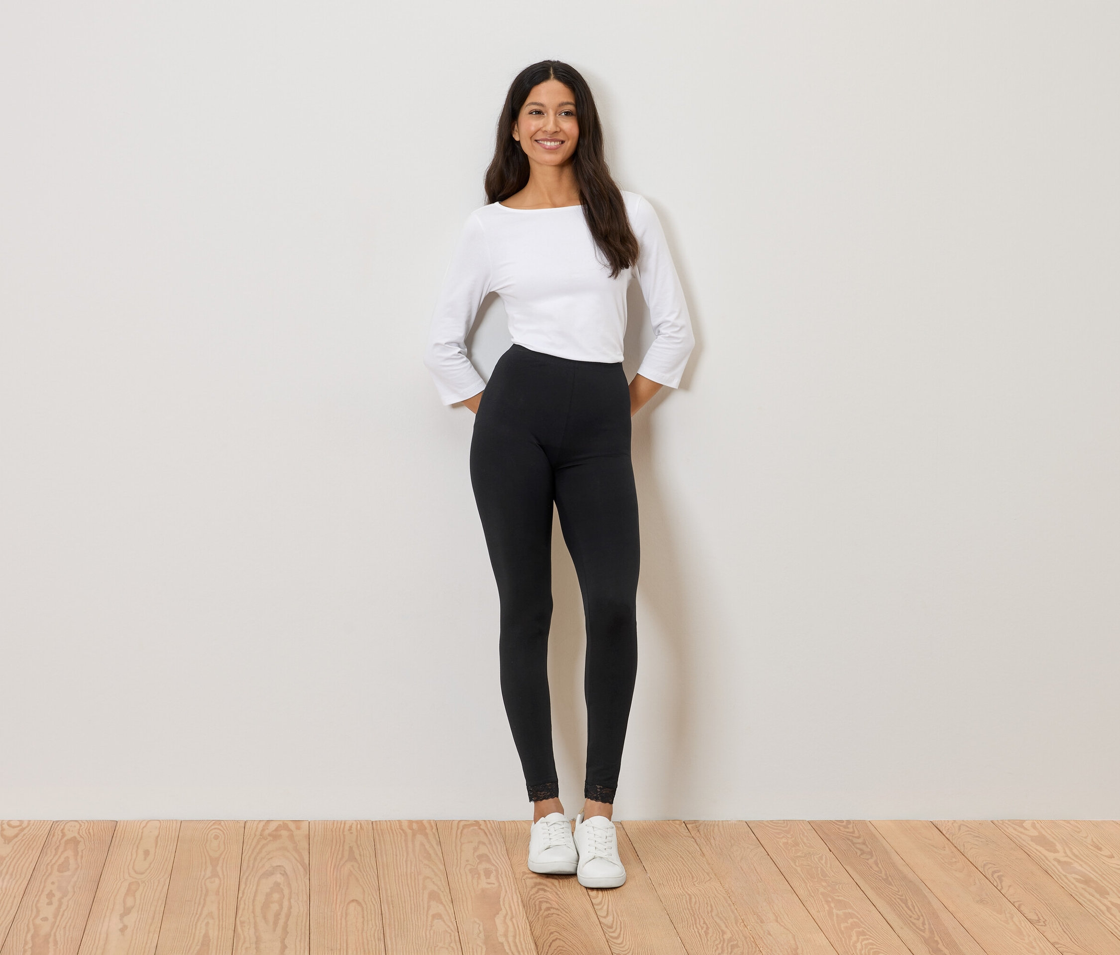 Kobieta stoi na tle ściany, ubrana w białą bluzkę, czarne legginsy i białe tenisówki.