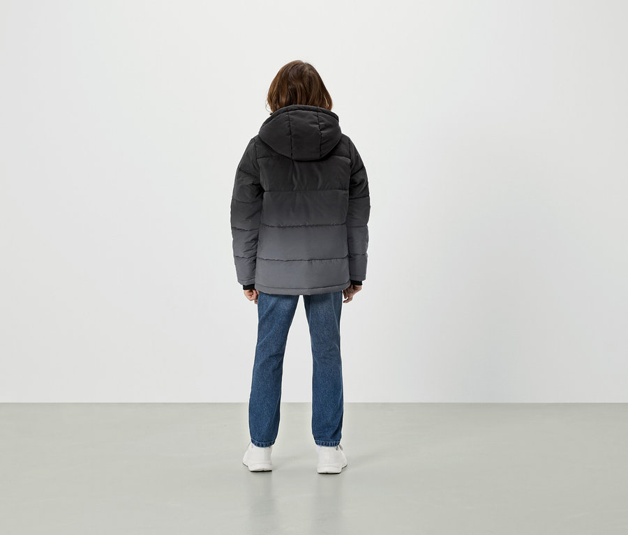 Widok z tyłu osoby w szarej puchowej kurtce z kapturem, niebieskich dżinsach i białych butach.