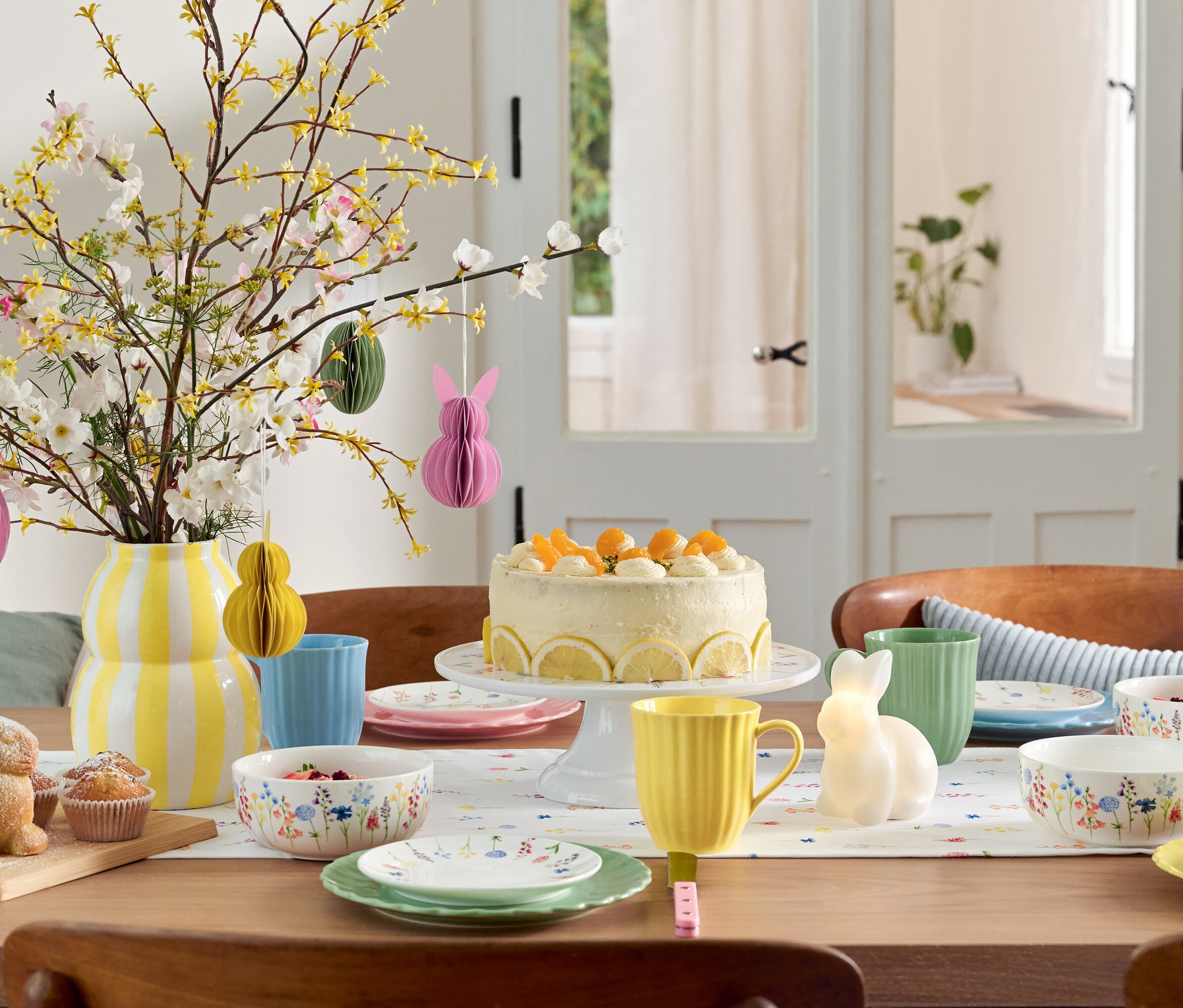 Stół wielkanocny z ciastem, naczyniami i wazonem z kwiatami.