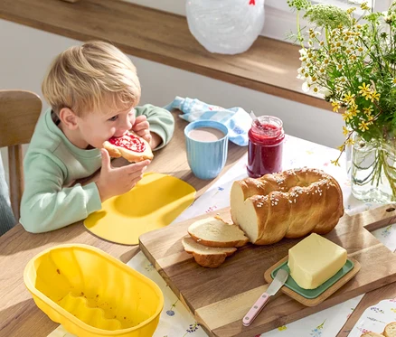 Blondwłosy chłopiec je kanapkę z dżemem przy stole z chałką, masłem, dżemem, naczyniami i kwiatami.