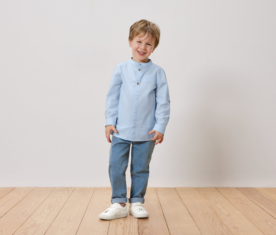 Uśmiechnięty blondwłosy chłopiec stoi na tle białej ściany. Ma na sobie jasnoniebieską koszulę, dżinsy i białe buty.