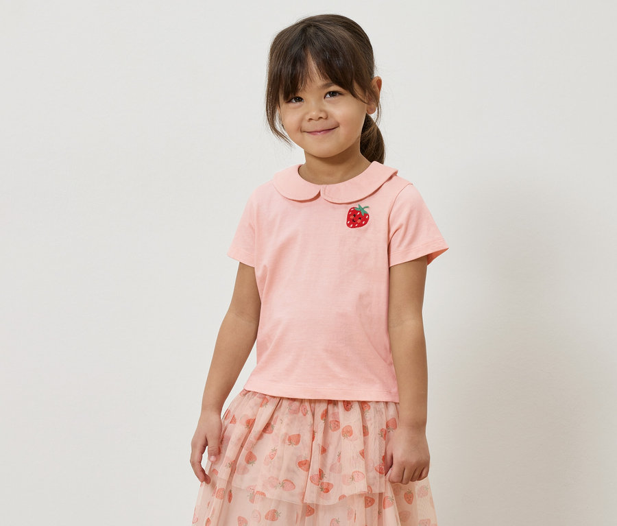 Dziewczynka w różowej koszulce z aplikacją truskawki i różowej spódnicy we wzór truskawek.