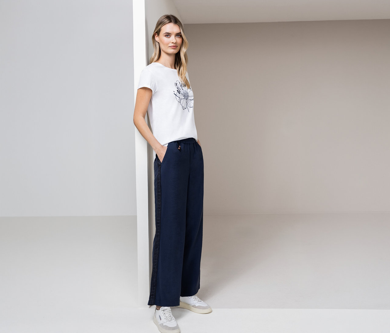 Kobieta pozuje w koszulce Street One, niebieskich spodniach i białych butach sportowych w jasnym pokoju.