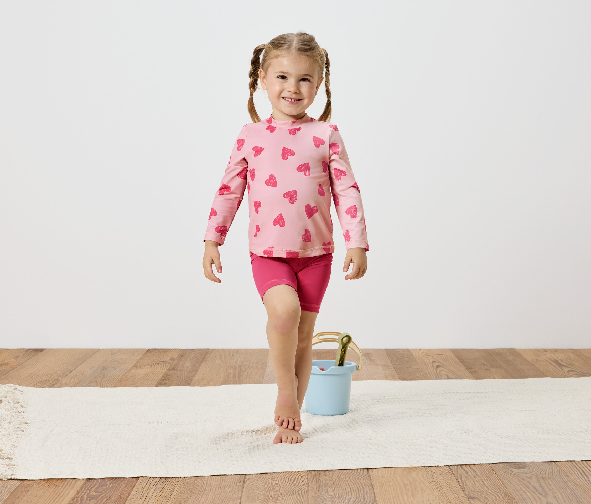 Dziewczynka w różowej bluzce w serduszka i spodenkach, balansuje na białym dywanie obok jasnoniebieskiego wiaderka.