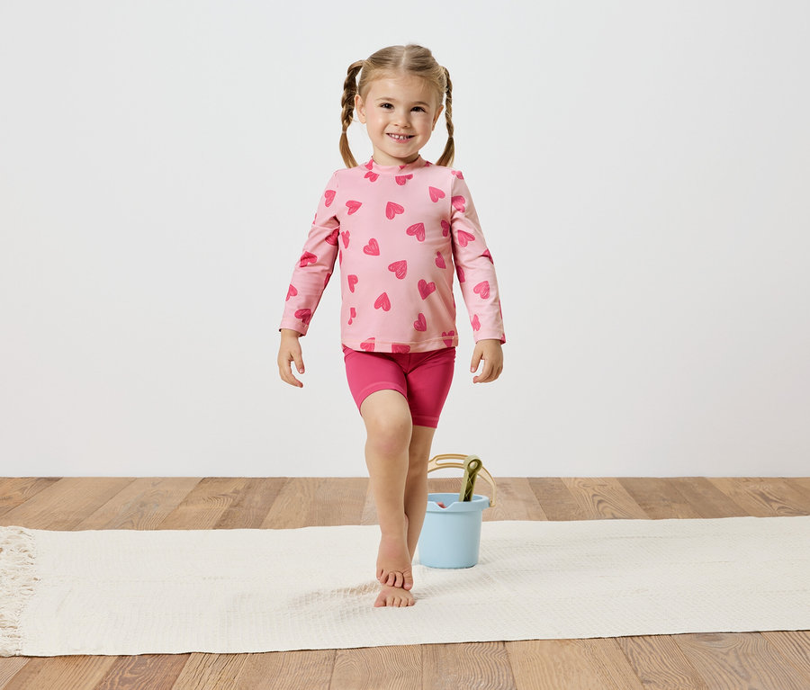 Dziewczynka w różowej bluzce w serduszka i spodenkach, balansuje na białym dywanie obok jasnoniebieskiego wiaderka.