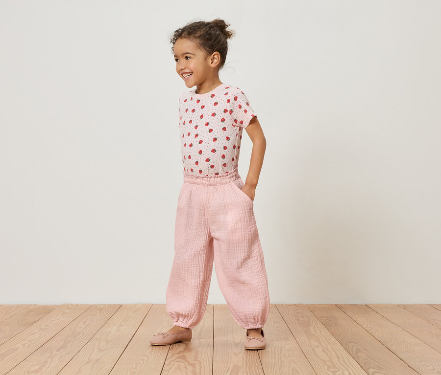 Dziewczynka stoi na drewnianej podłodze w różowej koszulce w truskawki i różowych spodniach.