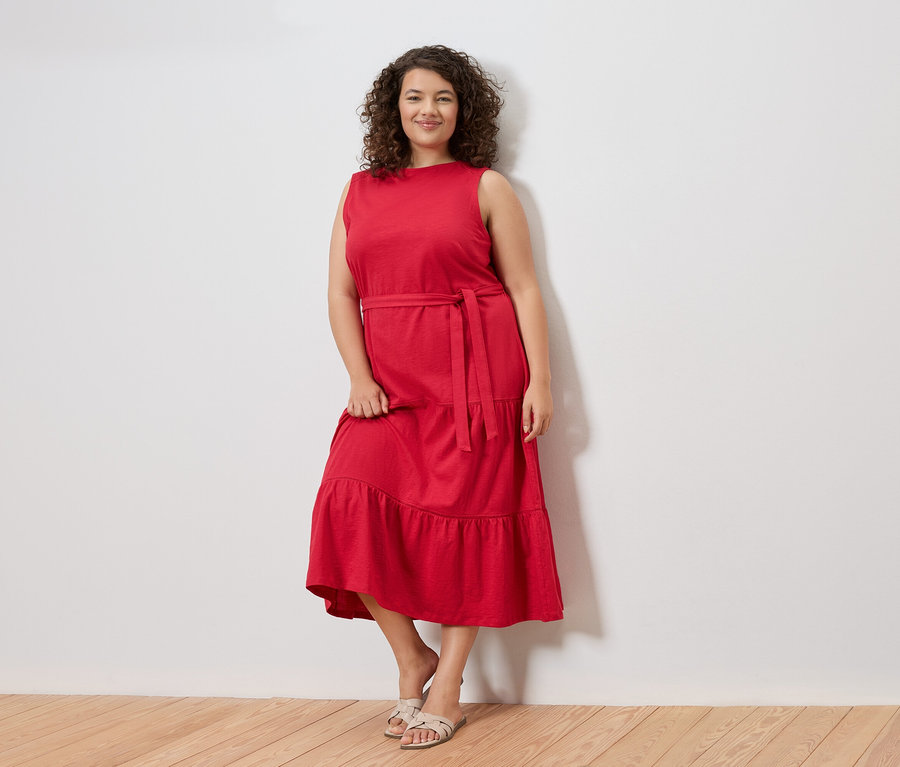 Kobieta o kręconych włosach stoi na tle białej ściany, ubrana w czerwoną sukienkę bez rękawów z paskiem i sandały.