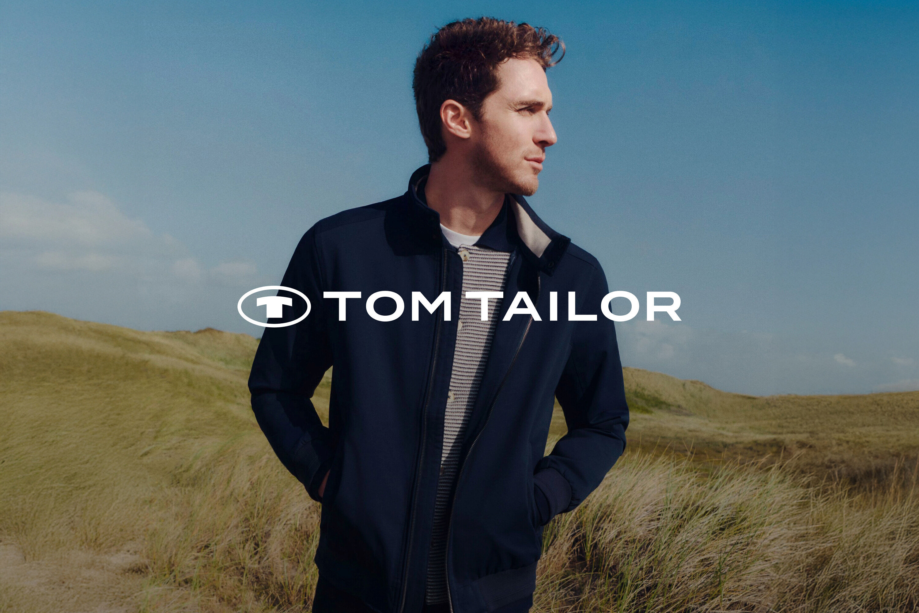 Mężczyzna w granatowej kurtce i koszuli w paski na tle trawiastego krajobrazu i błękitnego nieba.