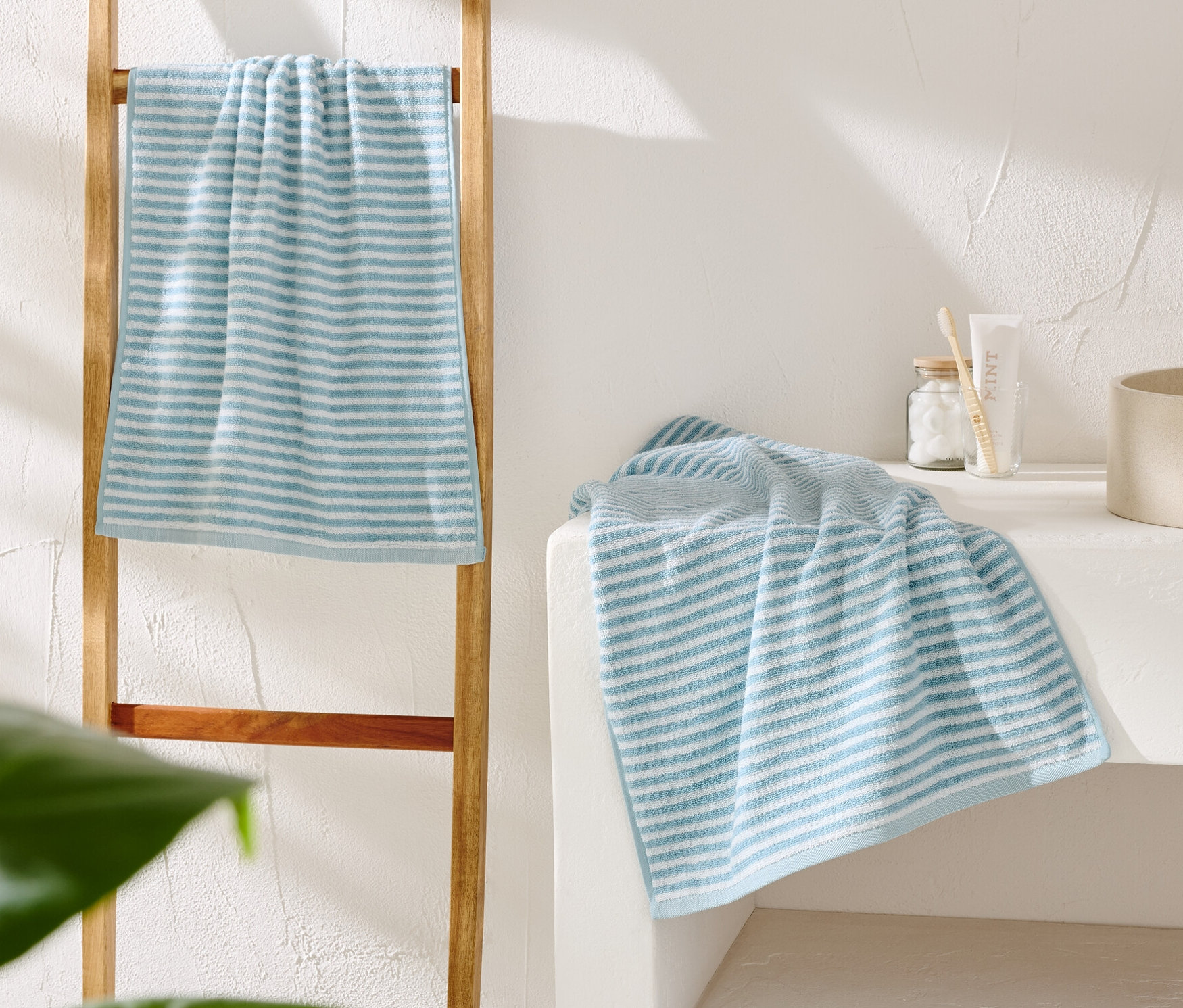 Pasiasty, biało-niebieski ręcznik zwisa z drewnianej drabiny. Obok beżowej umywalki leży drugi ręcznik.