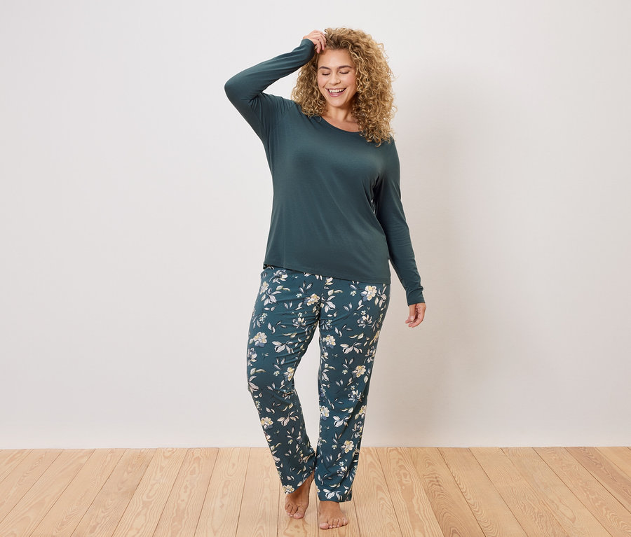 Kobieta z kręconymi włosami ma na sobie ciemnozieloną bluzkę z długim rękawem i ciemnozielone spodnie od piżamy w kwiatowy wzór.