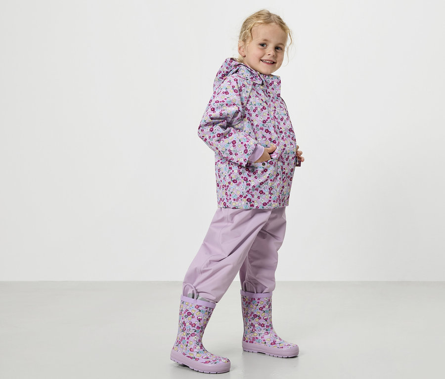 Blondwłosa dziewczynka w liliowym płaszczu przeciwdeszczowym w kwiaty, liliowych spodniach przeciwdeszczowych i pasujących kaloszach.