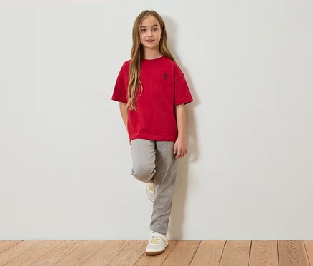 Dziewczyna z długimi brązowymi włosami, w czerwonym T-shircie i szarych spodniach dresowych, oparta o białą ścianę.