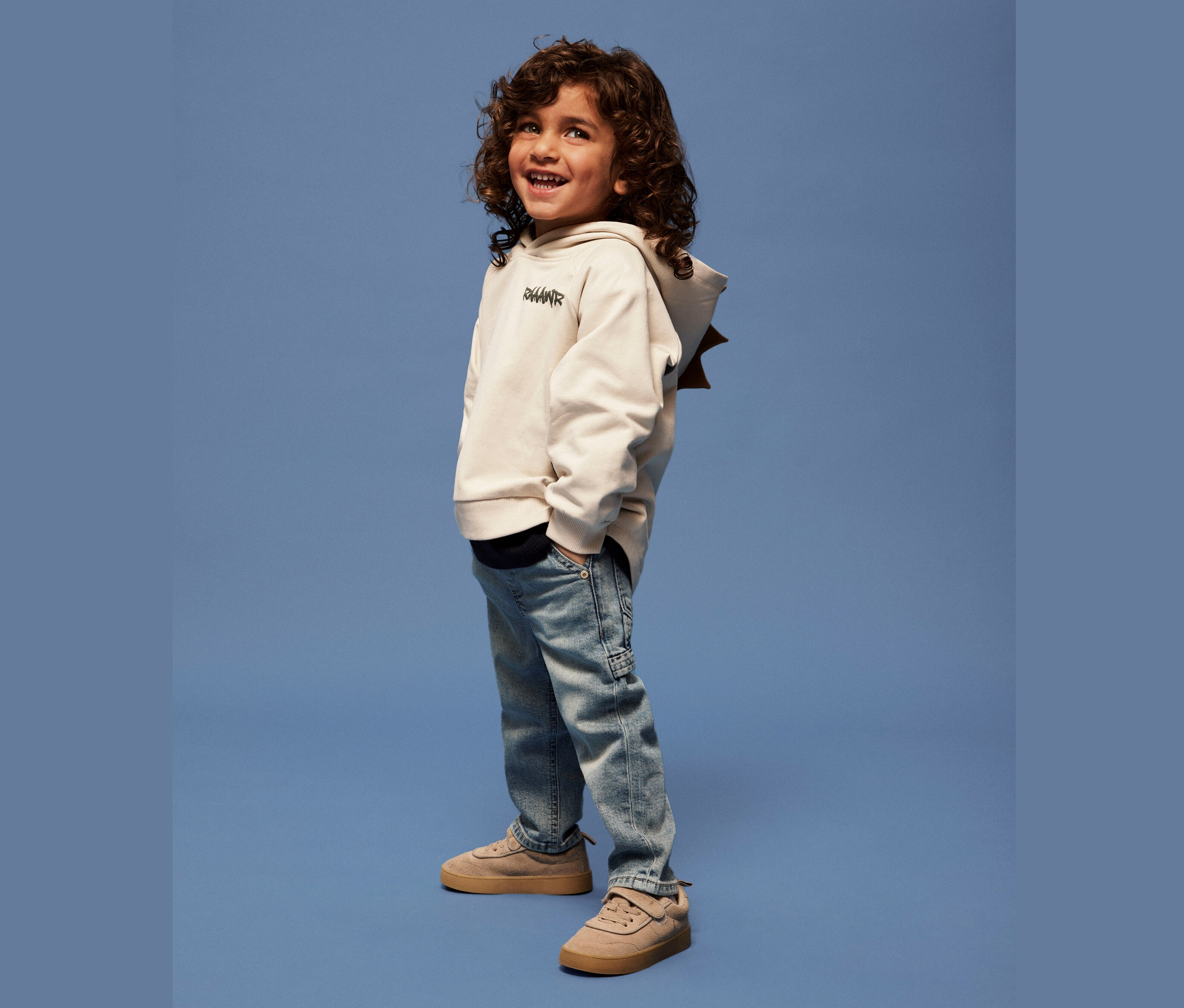 Uśmiechnięty chłopiec z kręconymi włosami stoi na niebieskim tle w beżowej bluzie z kapturem, jasnoniebieskich dżinsach i brązowych butach.