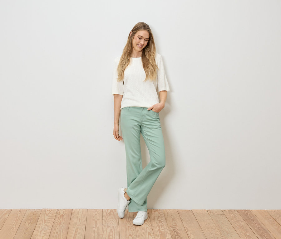 Kobieta o blond włosach w białym topie, zielonych spodniach i białych trampkach stoi na drewnianej podłodze na tle białej ściany.