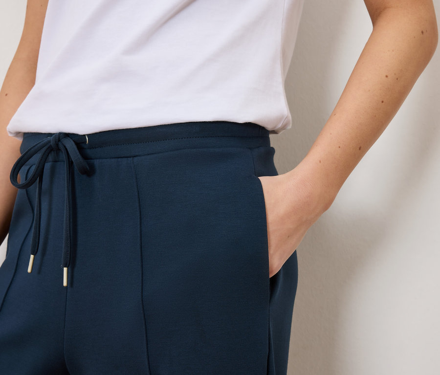 Zbliżenie na kobietę ubraną w granatowe spodnie dresowe ze sznurkiem i złotymi detalami oraz białą koszulkę.