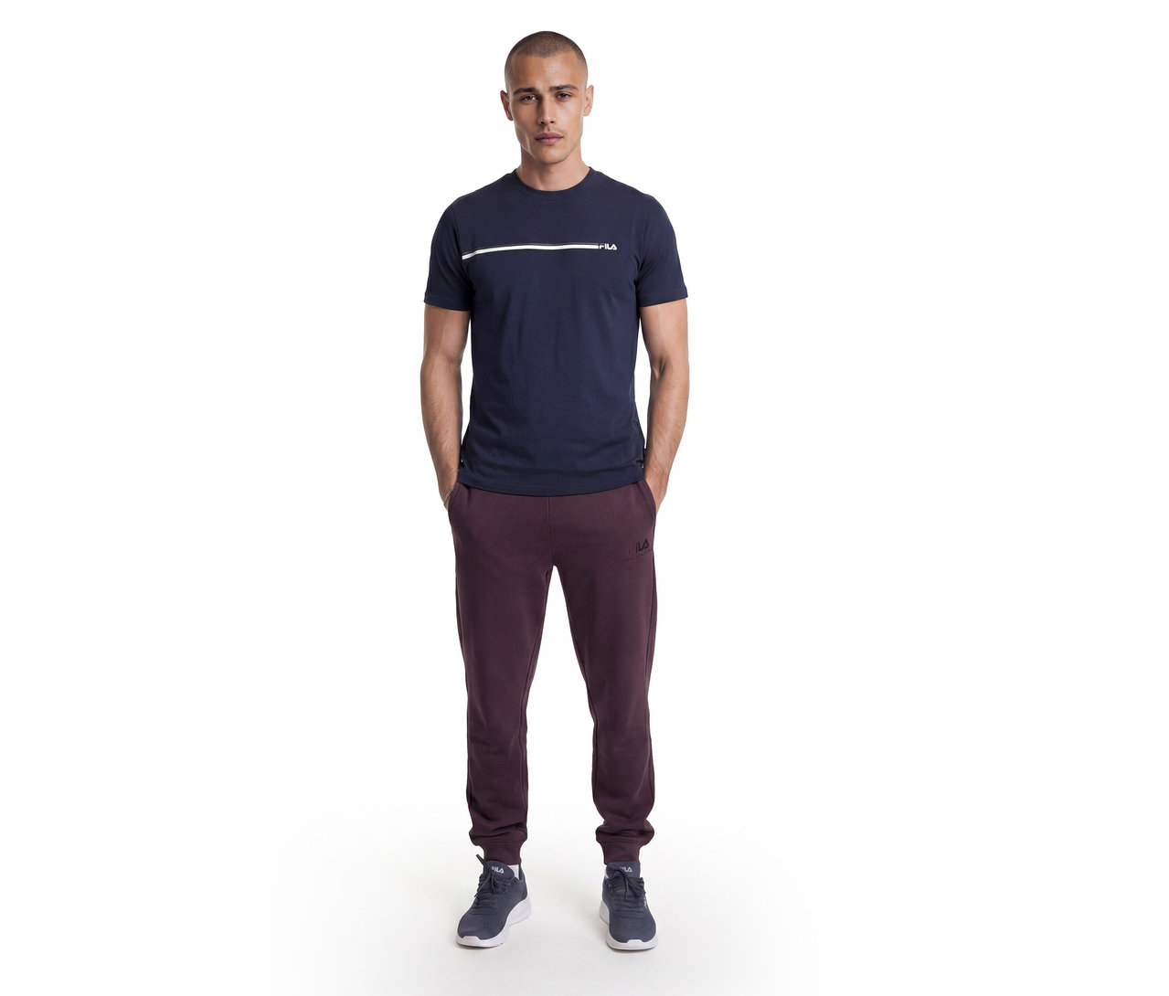 Mężczyzna stoi z rękami w kieszeniach, ubrany w granatowy T-shirt z białym paskiem i logo Fila, bordowe spodnie dresowe Fila oraz ciemnoniebieskie sneakersy.