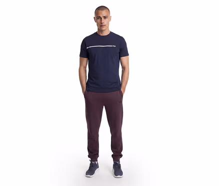 Mężczyzna stoi z rękami w kieszeniach, ubrany w granatowy T-shirt z białym paskiem i logo Fila, bordowe spodnie dresowe Fila oraz ciemnoniebieskie sneakersy.
