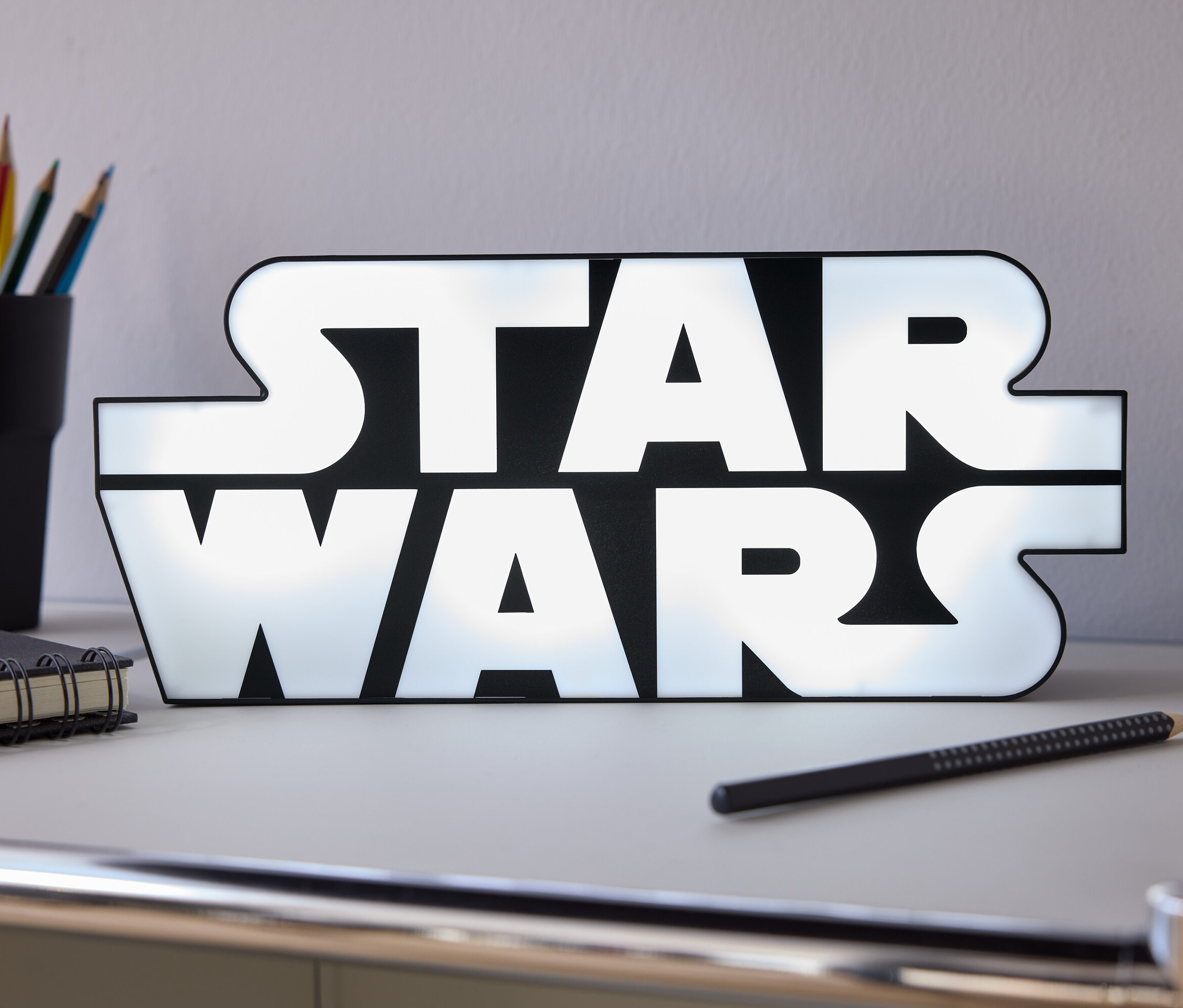 Na stole stoi świecący napis Star Wars, obok niego leży ołówek, notes i kubek z kredkami.