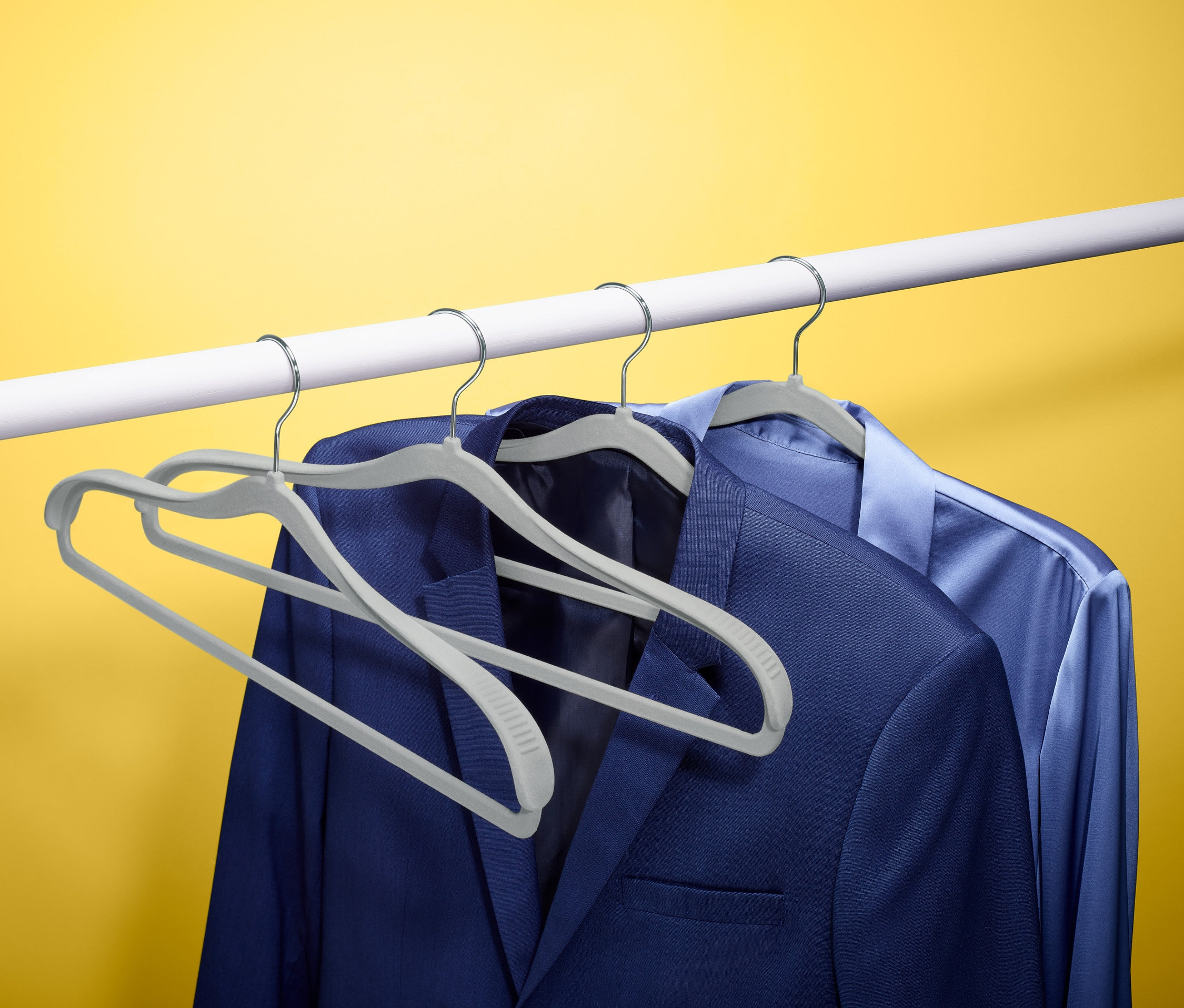 Dwa z czterech wieszaków na ubrania trzymają niebieską marynarkę i koszulę na białym drążku na tle żółtego tła.