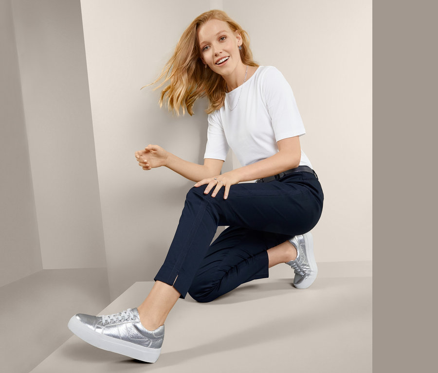 Kobieta siedzi na schodach i pozuje w damskich spodniach o długości do kostek z lampasami, białym t-shircie i srebrnych butach.