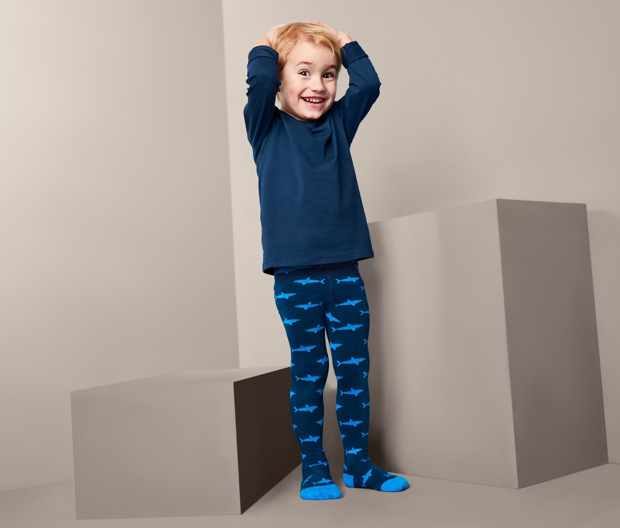 Chłopiec w niebieskiej koszulce i rajstopach we wzór rekinów stoi między szarymi blokami. Dziecięce rajstopy, 3 pary.