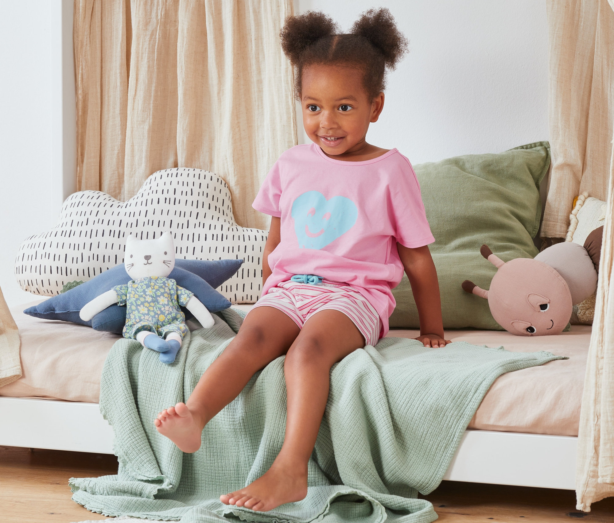 Dziewczynka siedzi na łóżku w piżamie z koszulką i spodenkami z bawełny ekologicznej, 2 sztuki.