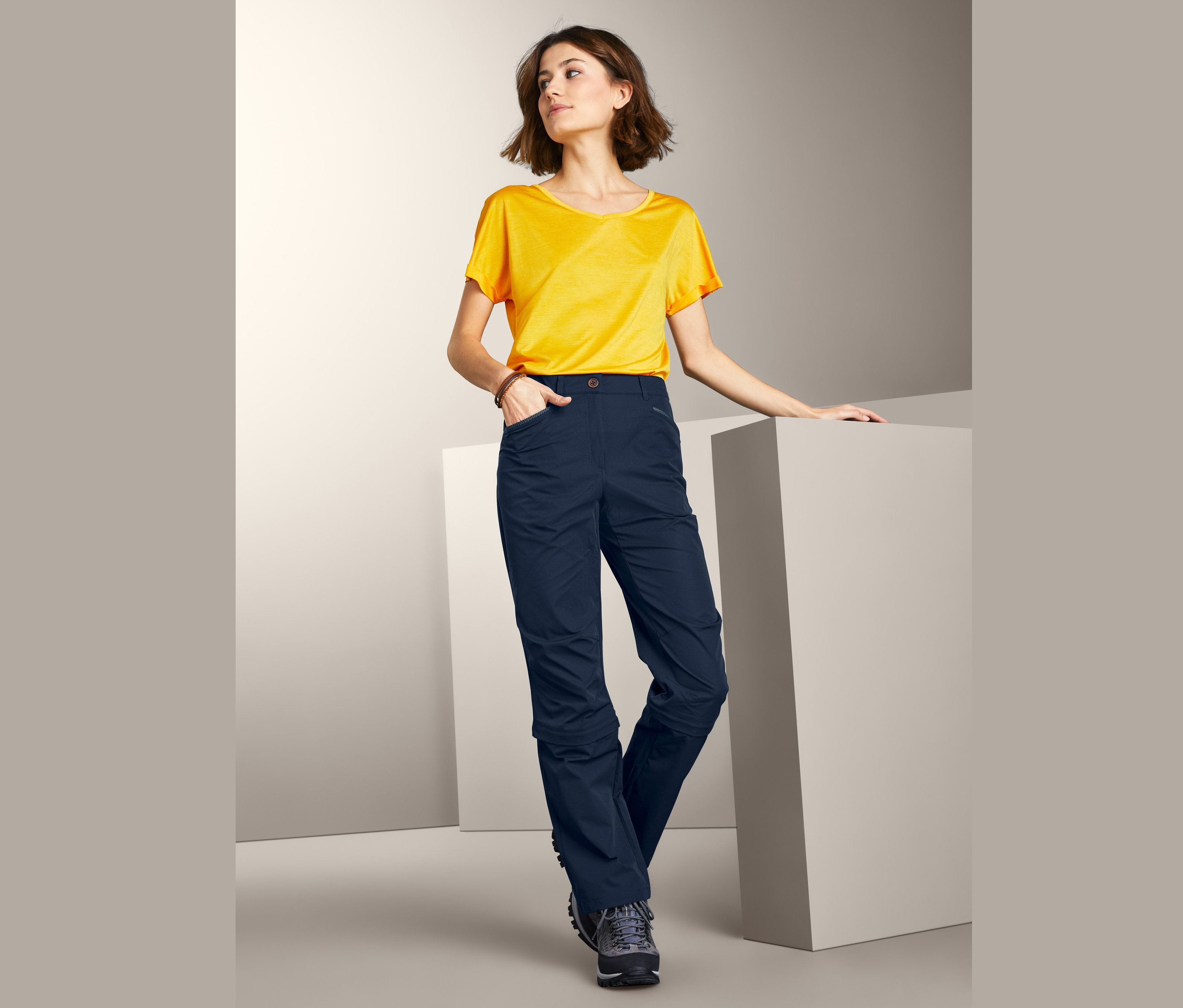 Kobieta pozuje w granatowych spodniach funkcyjnych z odpinanymi nogawkami i żółtej koszulce.