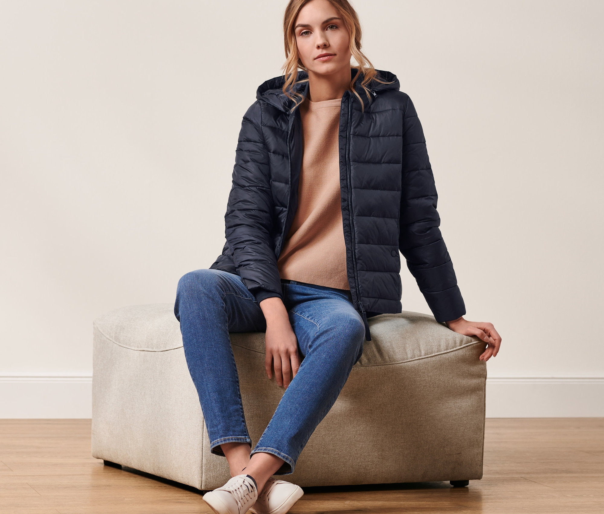 Kobieta siedzi na pufie i ma na sobie granatową kurtkę pikowaną z kapturem.
