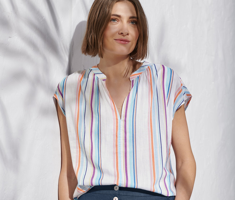 Kobieta oparta o ścianę ma na sobie tkaną bluzkę w paski i granatowe spodnie.