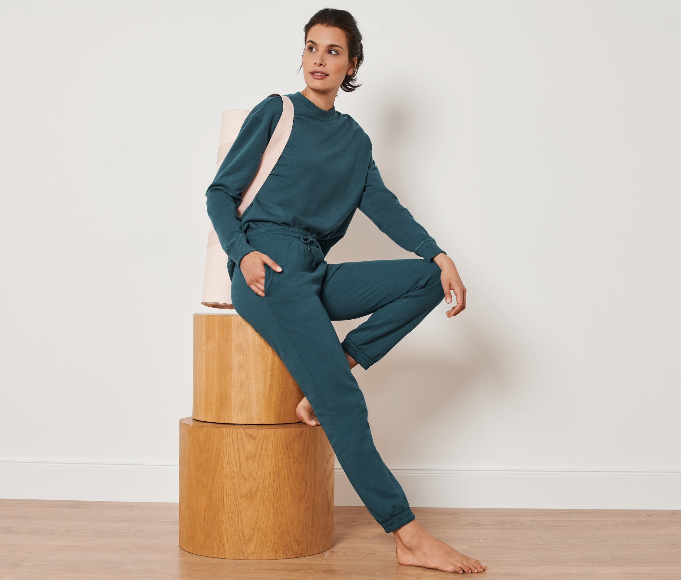 Kobieta siedzi na drewnianych taboretach, ubrana w spodnie dresowe w kolorze ciemnej zieleni szmaragdowej.