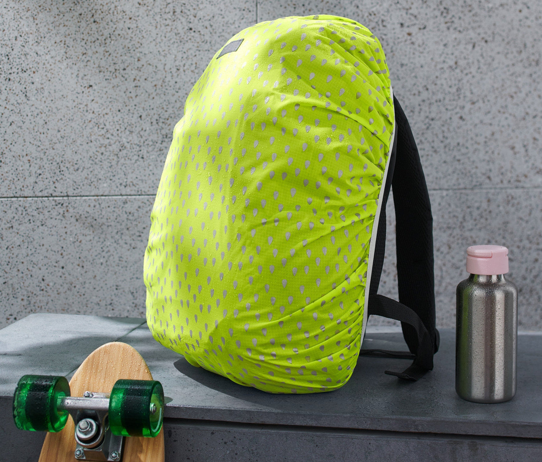 Plecak z odblaskowym pokrowcem przeciwdeszczowym, obok deskorolka i butelka na wodę.