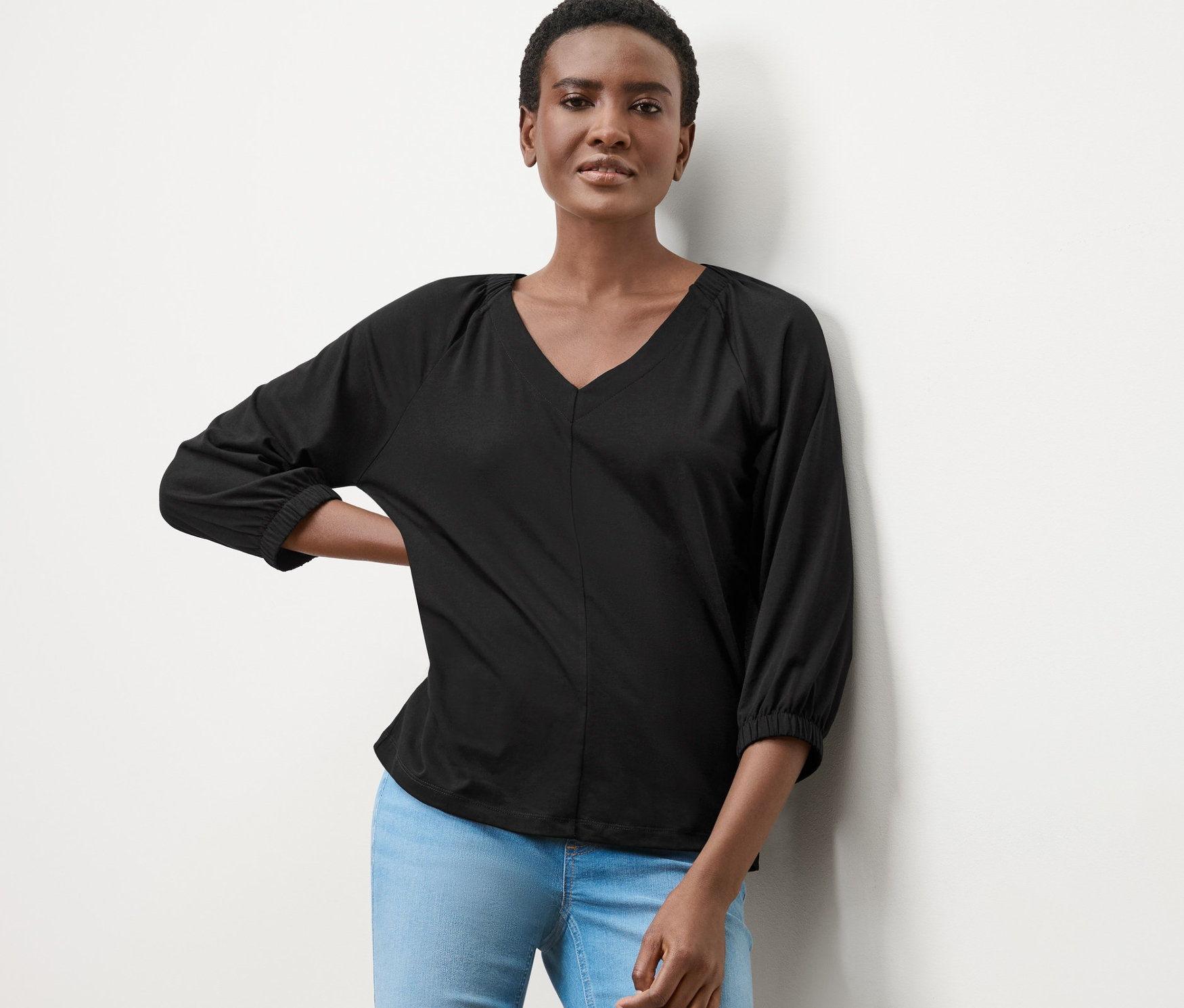 Kobieta pozuje w czarnej bluzce z marszczeniem i jasnych dżinsach.