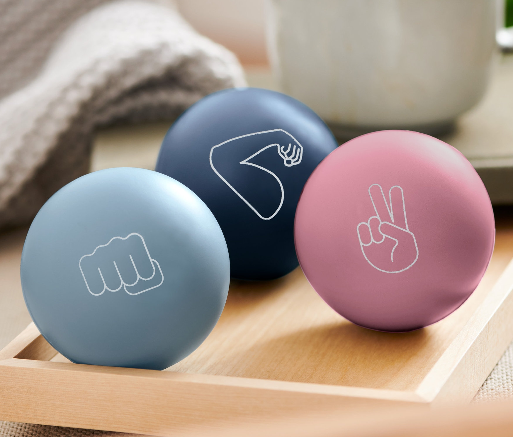 Na drewnianej tacy leżą trzy piłki antystresowe: jasnoniebieska z pięścią, ciemnoniebieska z ramieniem i różowa ze znakiem pokoju. W tle kubek.