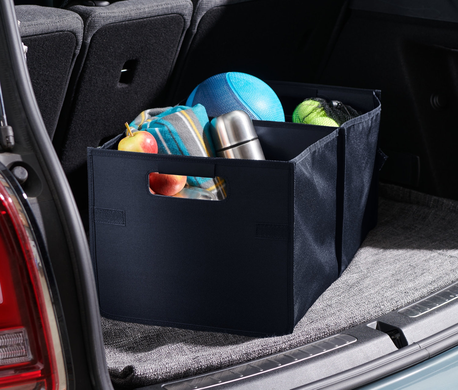 Składane pudło do samochodu leży w bagażniku samochodu. W pudełku leżą koc, piłka i termos.