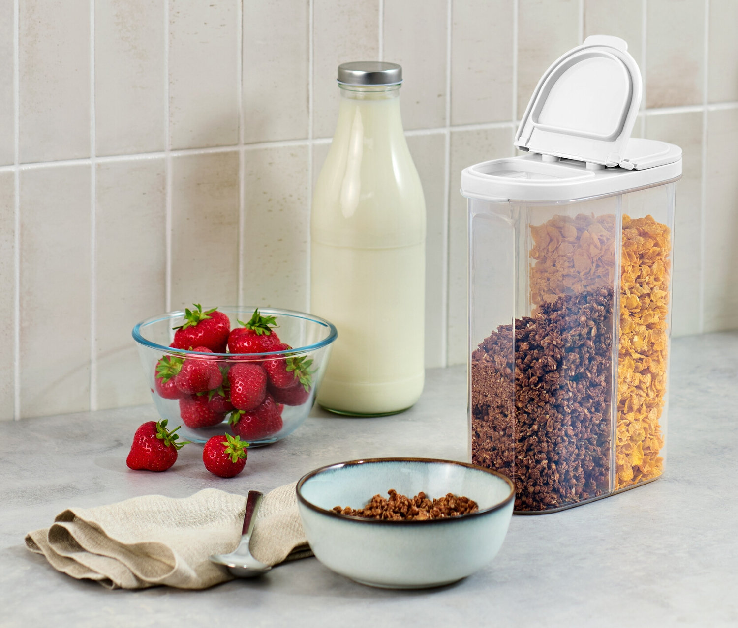 Na blacie kuchennym stoi pojemnik na żywność 2 w 1, miska truskawek, butelka mleka i miska płatków śniadaniowych.