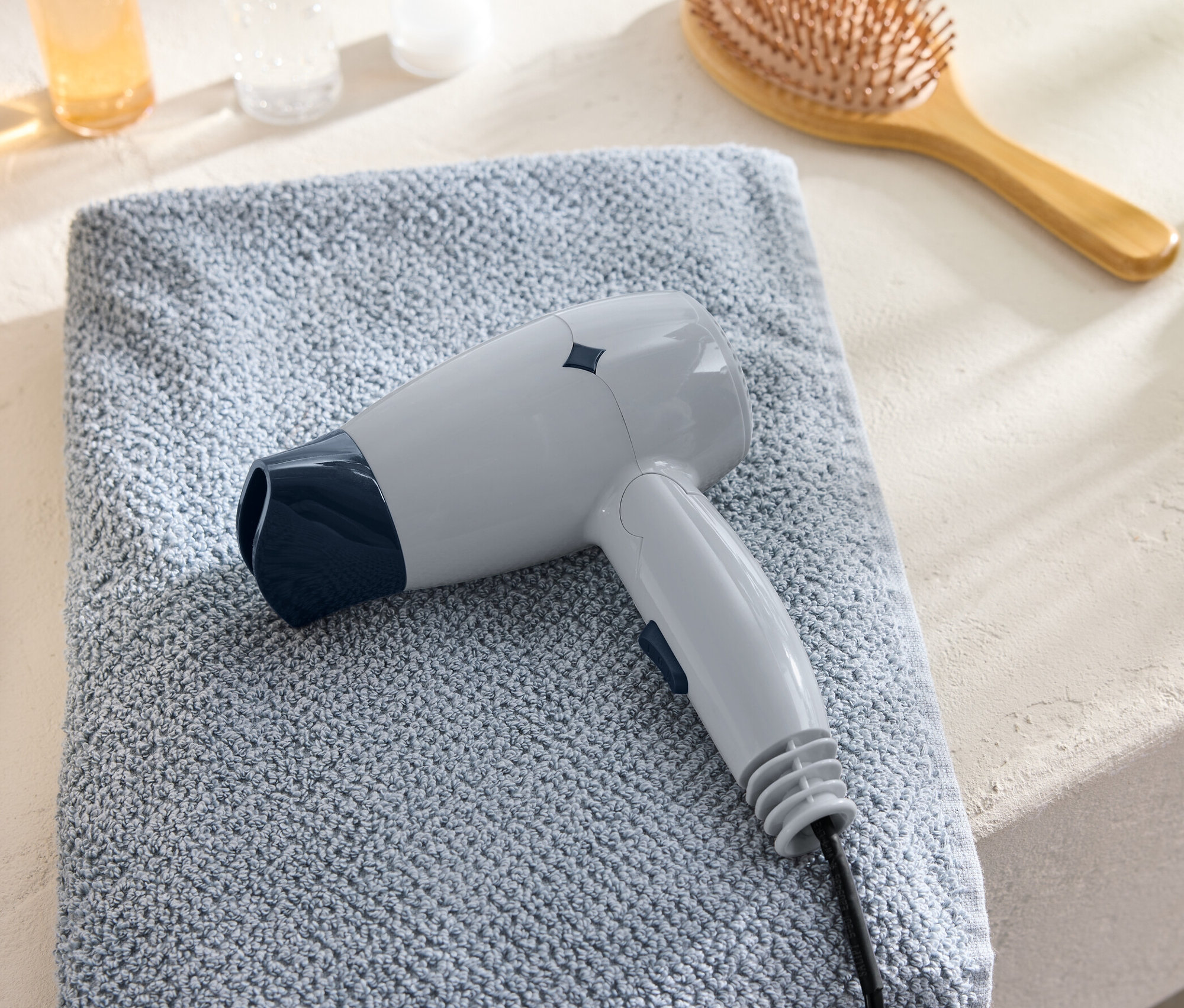 Szara podróżna suszarka do włosów leży na niebieskim ręczniku obok szczotki do włosów.
