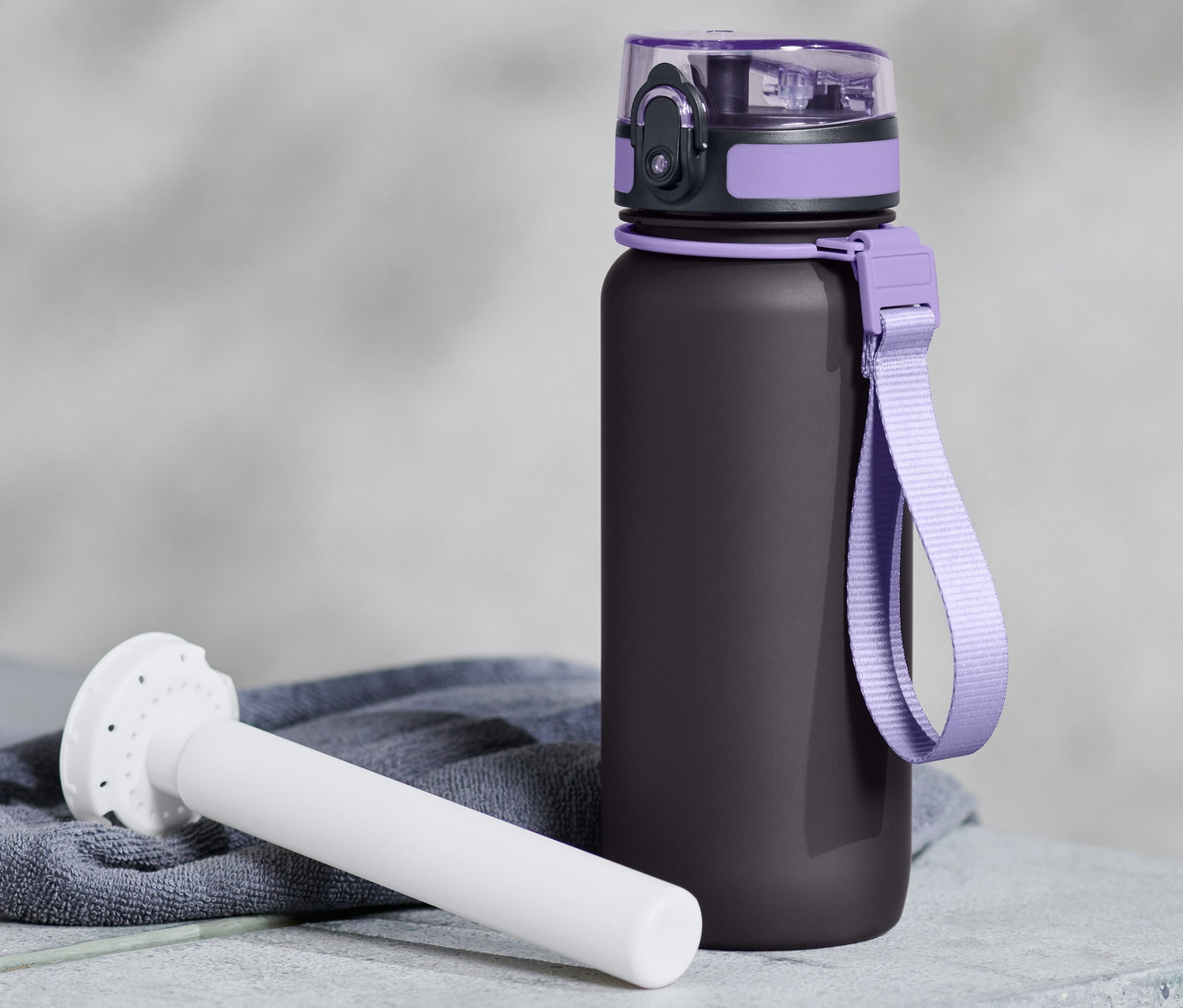 Czarna bidon z fioletowymi detalami i białym elementem chłodzącym leżą na szarej powierzchni.