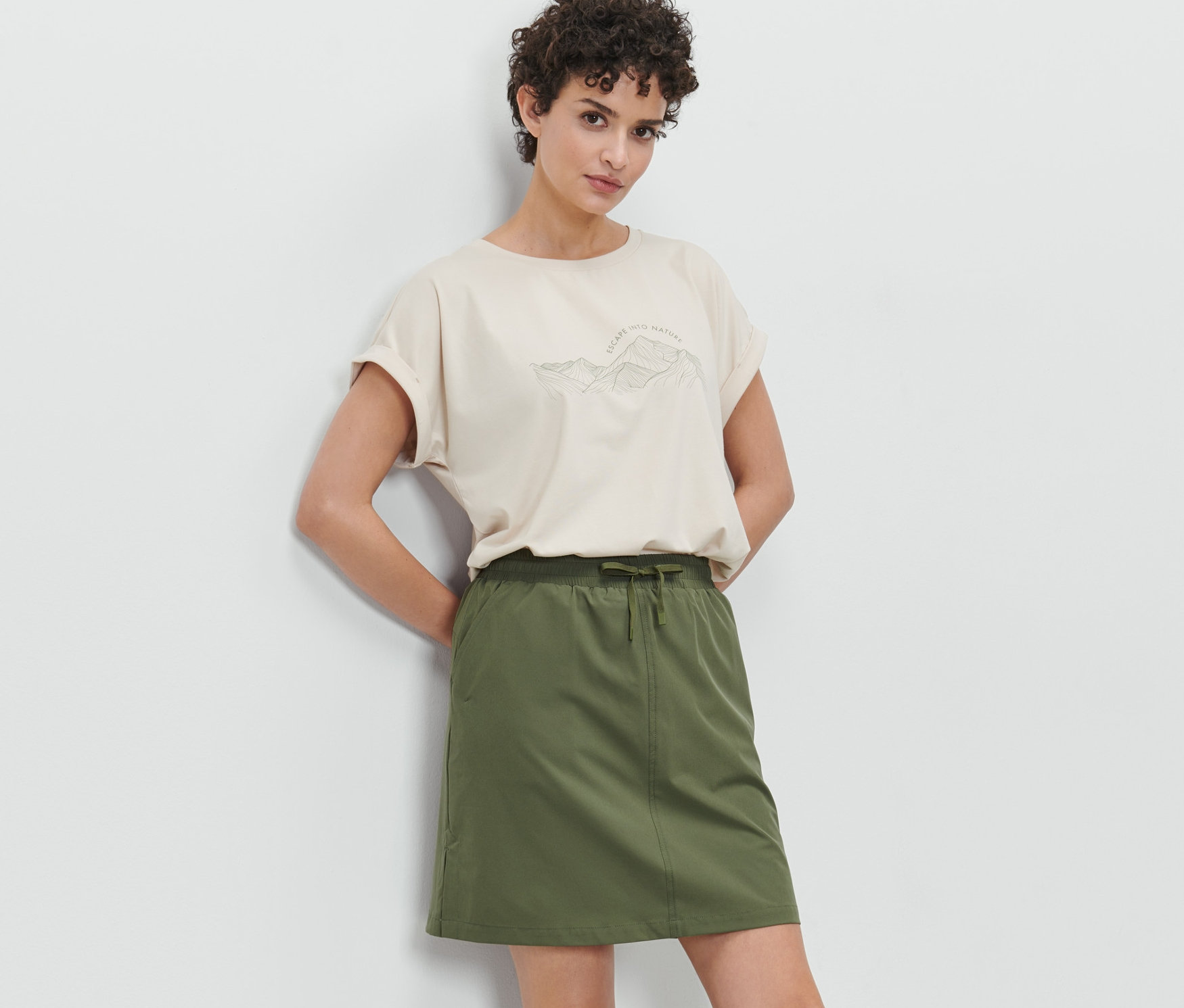 Kobieta pozuje w oliwkowej spódnicy funkcyjnej z krótkimi spodenkami i beżowym T-shircie.