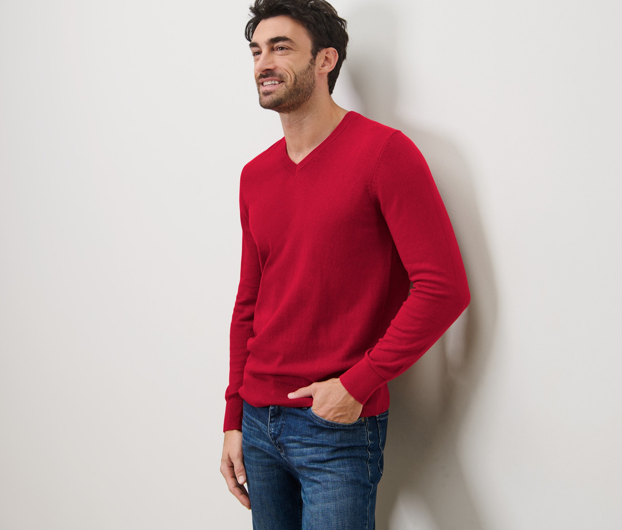 Uśmiechnięty mężczyzna opiera się o ścianę, ubrany w czerwony sweter z dekoltem w serek i dżinsy.