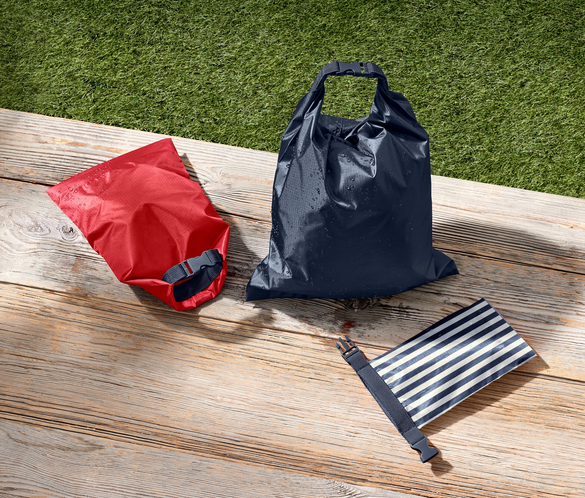 Czerwony, granatowy i niebiesko-biały w paski wodoszczelny worek leżą na drewnianej powierzchni obok zielonej trawy.