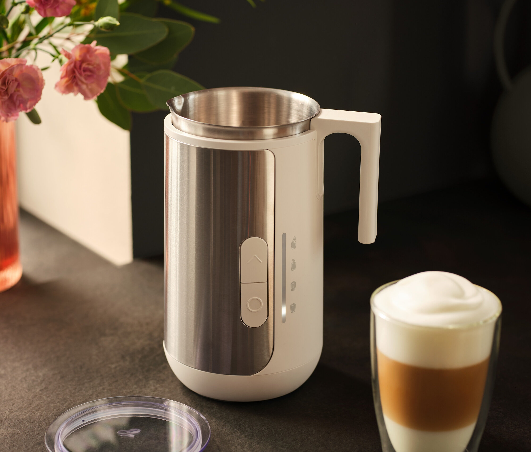 Spieniacz do mleka ze stali nierdzewnej stoi obok szklanki kawy latte i pokrywki na ciemnym blacie.