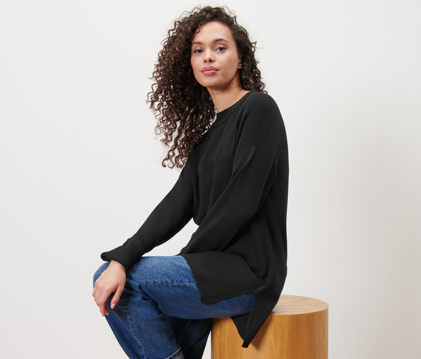 Kobieta siedzi na drewnianym stołku, ubrana w długi czarny sweter z dzianiny.