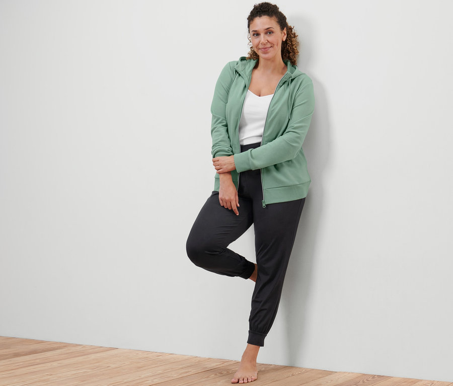 Kobieta opiera się o ścianę, ubrana w bluzę z dzianiny dresowej w kolorze szałwiowej zieleni i spodnie do jogi.