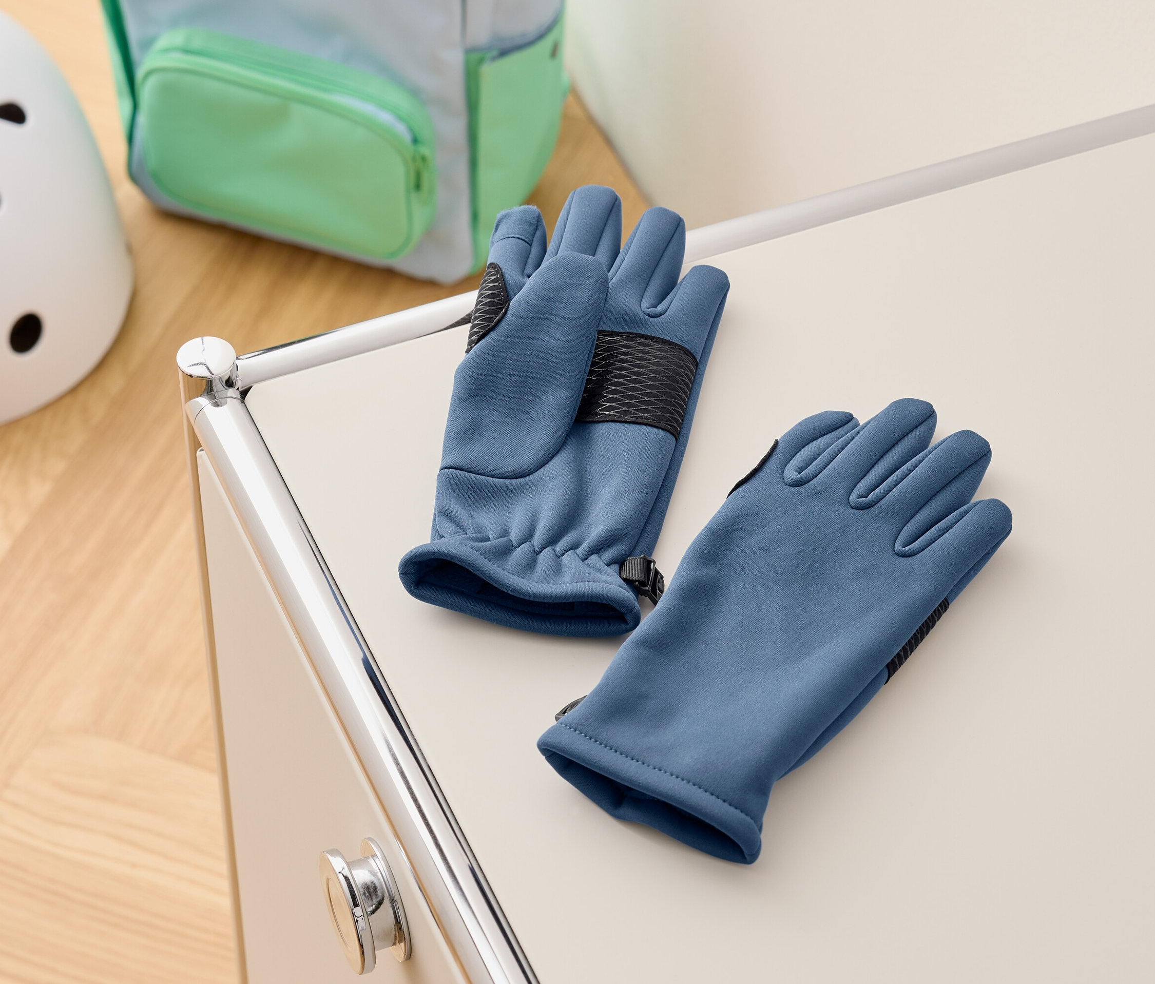 Niebieskie rękawice leżą na białej szafce, w tle plecak i kask.