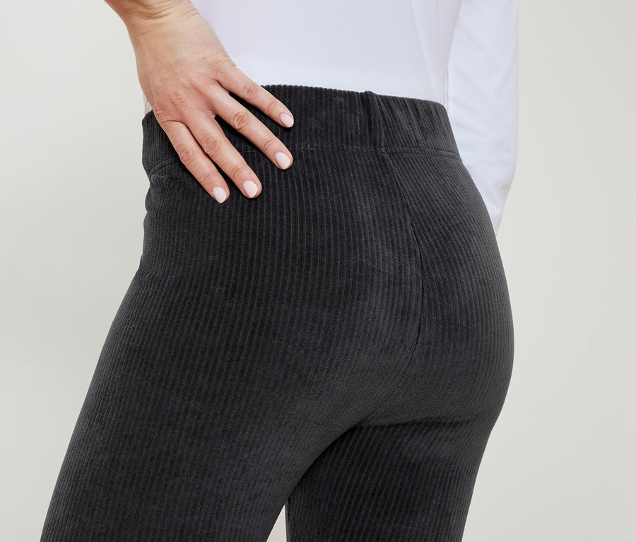 Kobieta w szarych spodniach sztruksowych kładzie rękę na biodrze.