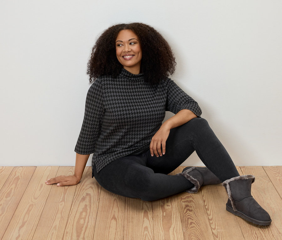 Kobieta siedzi na podłodze, ma na sobie sztruksowe legginsy, wzorzystą bluzkę i buty.